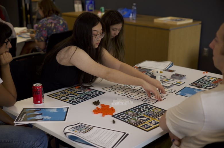 A Tisch Summer High School Game Design student seated at a table reaches out to turn over one of the cards arranged on the table. Other students sit and watch the student take her turn.